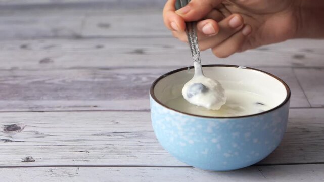 Person stirring fresh yogurt with blueberries in blue bowl on wooden background in cinematic video