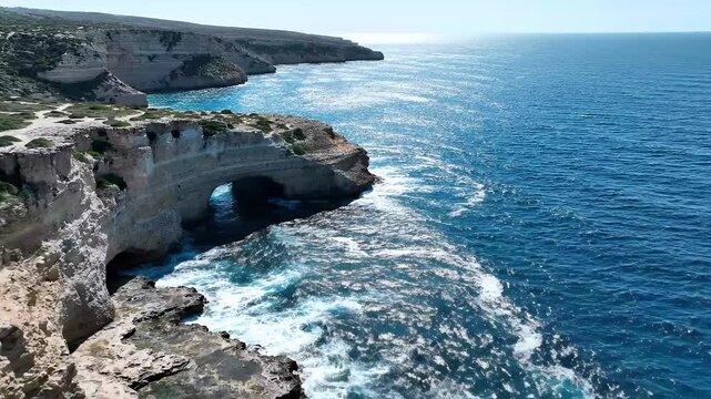 Sunlit ocean waves crash against rugged coastal cliffs with a natural archway, sparkling blue sea footage