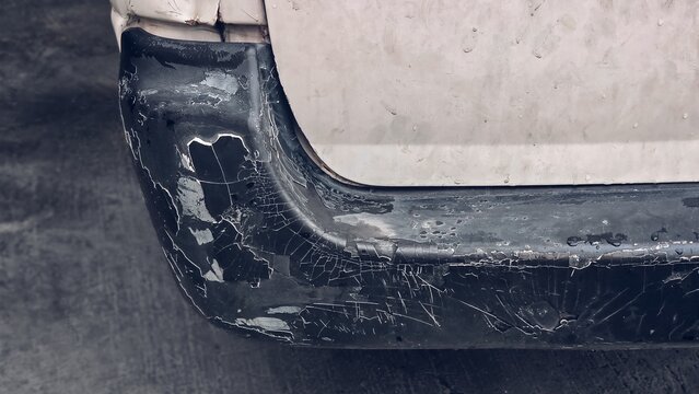 Close up of a heavily damaged black plastic car bumper with severely cracked, peeling, and flaking paint, showing deep scratches and weathered texture on a white vehicle.
