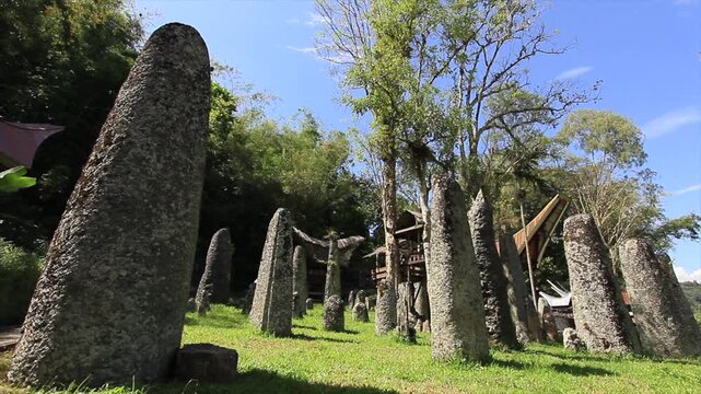 Ancient megalithic stone pillars (menhirs) standing in Bori Kalimpenne burial site under bright sunny sky with traditional Toraja Tongkonan house and lush green surroundings, South Sulawesi, Indonesia