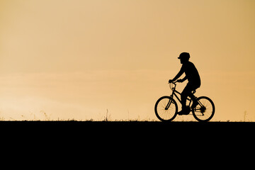 Obraz premium Side view silhouette of a cyclist wearing a helmet riding a bicycle during a warm sunset symbolizing safety active mobility outdoor exercise fitness routines and peaceful evening recreation.