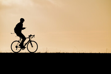 Obraz premium Distinct silhouette of a cyclist riding a bike while looking at a smartphone screen during golden hour sunset representing connected technology mobile communication active lifestyle and multitasking