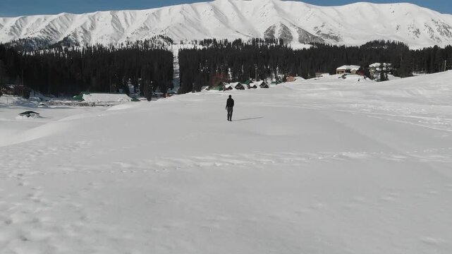 Solo traveller walking across snowy Gulmarg valley with Himalayan mountains and forest cabins in winter, Kashmir India tourism.