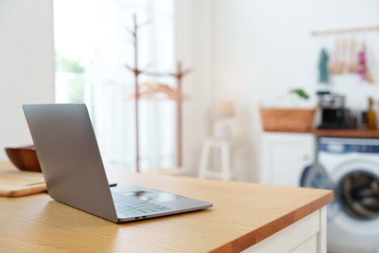 Modern computer laptop on wooden kitchen island table with washing machine counter in background in white cozy laundry room at home in sunlight from sheer curtain window
