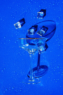 Empty cocktail glass with ice cubes on a wet, vibrant blue summer background.