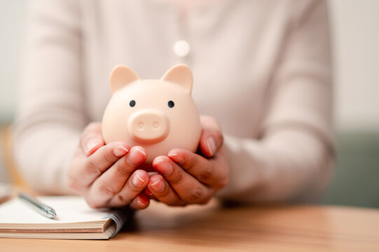 Financial security and saving money concept. Woman hands protecting a piggy bank on wooden desk with notebook.
