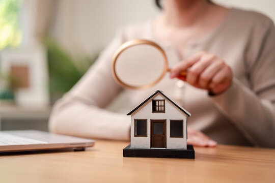Online property search and house hunting concept. Blurred woman hand holding magnifying glass near model house and laptop.