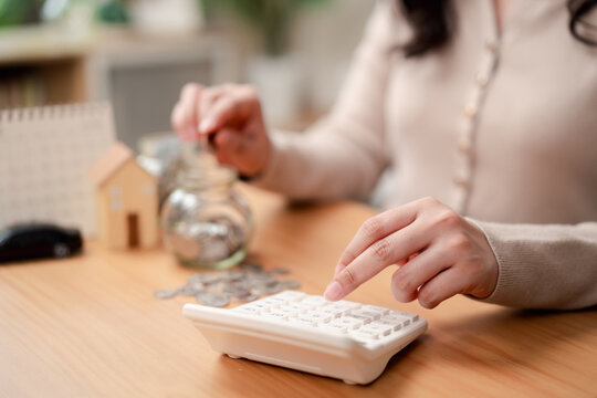Personal finance and saving goals concept. Woman hand dropping a coin into a glass jar with house and car models on desk.