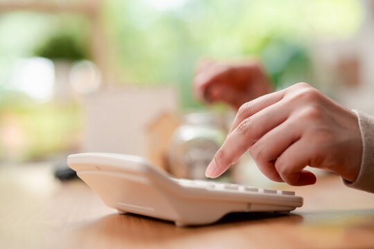 Personal finance and saving goals concept. Woman hand dropping a coin into a glass jar with house and car models on desk.