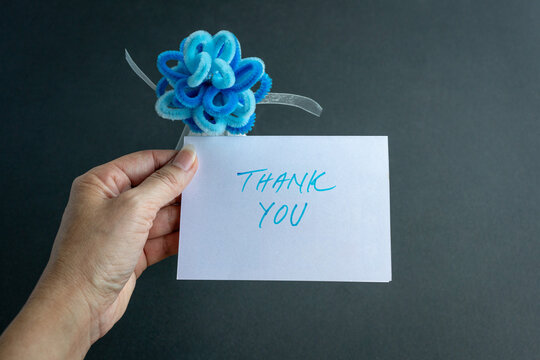 Hand holding a thank you card with blue artificial deco into flower, against black background.