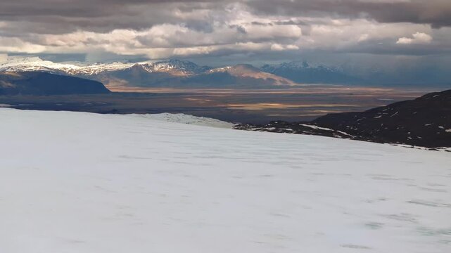 Snowy glacier plateau overlooking distant mountains beneath dramatic skies in Iceland, aerial overview of ice and desert