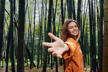 Fototapeta premium Woman in bamboo forest reaches hand toward camera, smiling in orange jacket. Portrait of young woman connecting with nature, walkway among tall green bamboo stalks.
