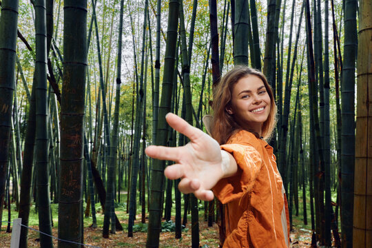 Woman in bamboo forest reaches hand toward camera, smiling in orange jacket. Portrait of young woman connecting with nature, walkway among tall green bamboo stalks.