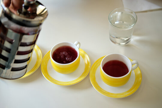 Two yellow teacups filled with hot herbal tea sit beside a stainless steel French press on a white table. Soft natural light and a glass of water create a inviting breakfast or afternoon tea scene.
