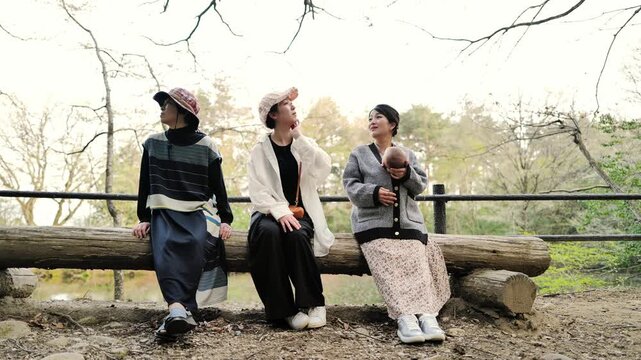 Three friends sit on a log bench outdoors. They look relaxed, enjoying each other's company. Branches frame the scene, suggesting a park or garden. The mood is calm and friendly