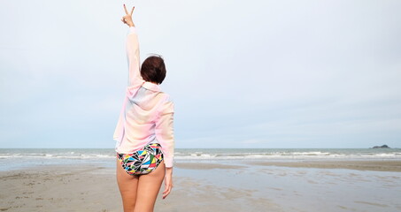 Woman in a colorful swimsuit and pastel cover up stands on the beach making a peace sign while facing the ocean © iFocus
