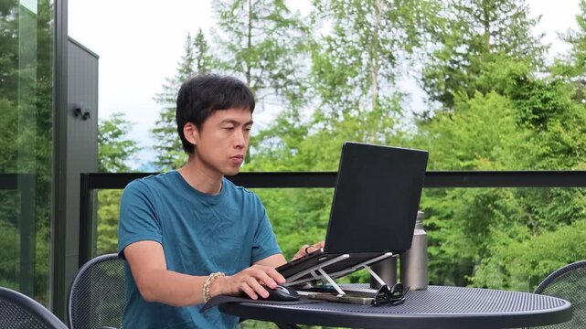 Young man works on laptop outdoors. Laptop rests on small stand. Green trees surround him. He types with focused expression. Perfect for remote work or study themes