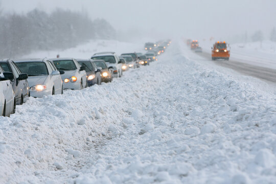 Snow blankets the road, cars paused in a long line. Foggy, gray skies obscure distant visibility and calm the scene. Drivers wait patiently, headlights casting soft glow