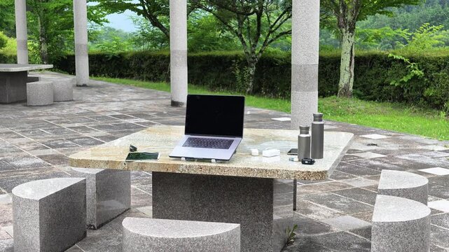 Modern stone table with laptop outdoors. Calm green surroundings enhance tranquil vibe. Laptop open, suggesting work or study. Natural light highlights stone texture and design