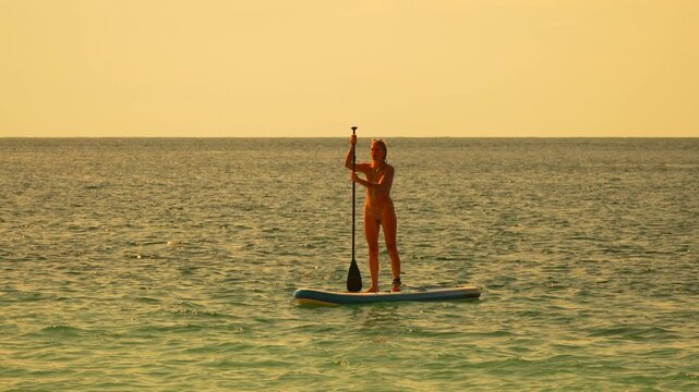 Paddleboarding woman sunset enjoys golden hour on the sea standing on a SUP board during summer vacation