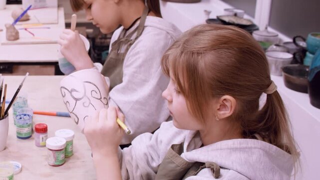 Master class. Artistic childhood. Two girls painting on cup using tool enjoying handmade activity in creative art artisan workshop.