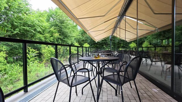 Outdoor cafe patio with modern furniture and greenery backdrop. Beige awning shades seating area under glass walls. Black metal chairs surround round tables on tiled floor