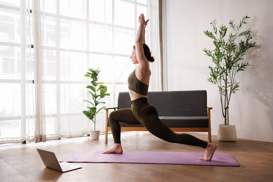 Woman practicing yoga at home, performing warrior pose in an online fitness class on a laptop, embracing wellness, mindfulness, and strength for mental and physical health
