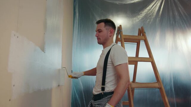 Handsome young man in a casual look actively paints the wall with a white paint. Wooden ladder, rubber sheet on the background. Positive mood, cheerful emotions. Slow motion, camera stabilizer shot