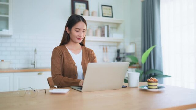 Beautiful young Asian woman working with laptop computer in home kitchen. Work at home