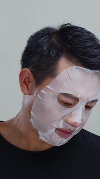 Asian man doing skincare treatment with sheet mask getting angry when answering call from boss. Businessman talking during morning routine.