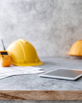 Yellow hard hats rest beside a tablet and walkie-talkie. Blueprints lie open on a rustic workbench. Modern tools meet classic safety gear in this scene