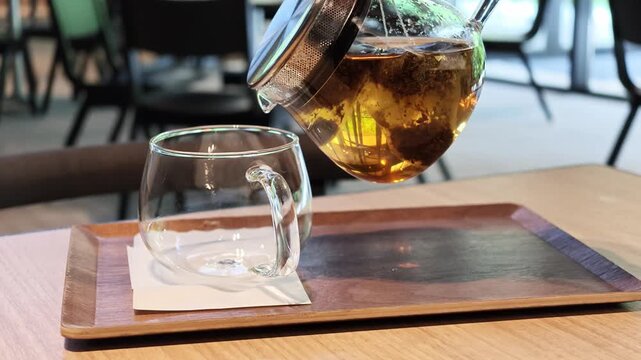 Warm tea pours from glass kettle into clear cup. Glass teapot rests beside filled glass cup. Wooden tray holds tea set on wooden table. Soft focus background shows cafe seating