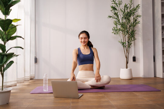 Asian woman doing yoga and meditation online at home, engaging in a healthy wellness routine and mindful fitness practice while using a laptop for virtual exercise