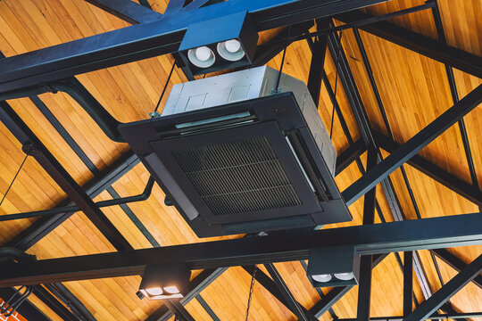 Modern cassette type air conditioner installed on loft ceiling with steel beams and wooden roof. Ceiling-mounted cassette air conditioning unit integrated into modern interior design. HVAC systems.