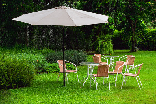 Garden patio set with white umbrella on lush green lawn. 
Outdoor seating area setup featuring woven wicker chairs, round tables, and large white patio parasol placed on vibrant green grass field.