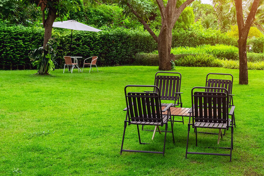 Outdoor patio seating sets on green lawn by river. Garden chairs and wooden tables on lush green grass field under shady trees in lakeside backyard, featuring peaceful riverfront landscape view.