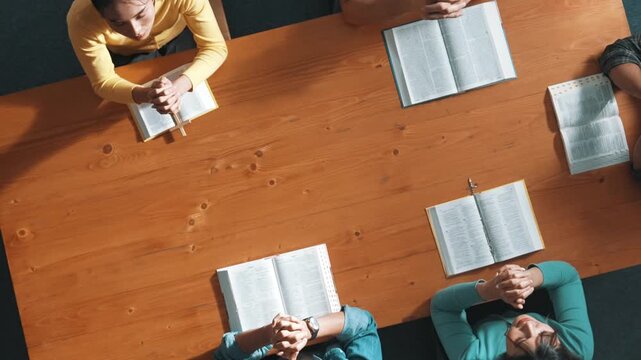 Top down aerial view of smart team hold hands on the holy bible and praying god while sitting at meeting table. Group of people hand folded in prayer with faith, spirituality and religion. Symposium.