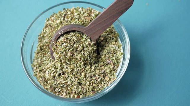Dried oregano herb in wooden spoon spilling from glass bowl on teal background