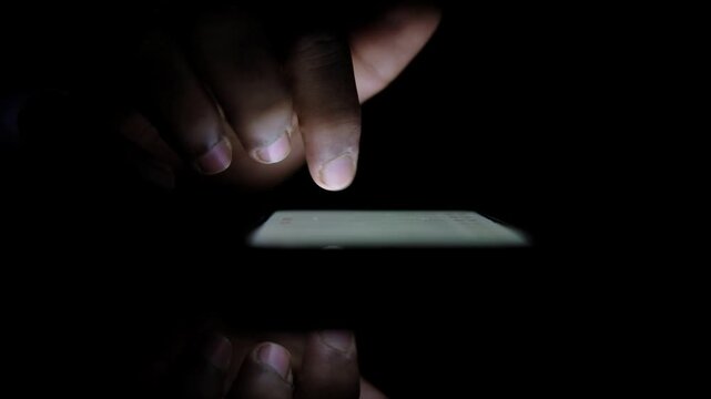 Close up slow motion of human finger touching typing scrolling smartphone screen in dark room at night with glowing screen reflection on glass table
