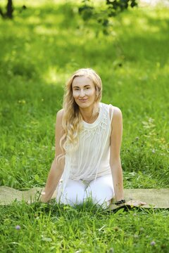 A woman with long hair sits on a yoga mat in a vibrant green park. She wears a light outfit and enjoys the peaceful surroundings. Sunlight filters through the trees, creating a serene atmosphere