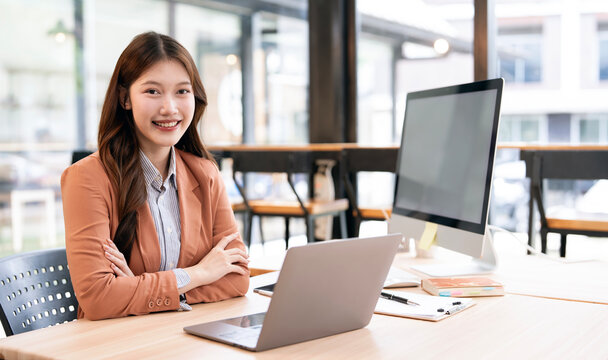 Portrait of confident young Asian businesswoman or entrepreneur smiling at desk in modern co-working space or office with laptop