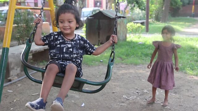 Indian little girl enjoying a playground swing while another girl gently pushes her in a public park, sharing a joyful outdoor childhood moment.