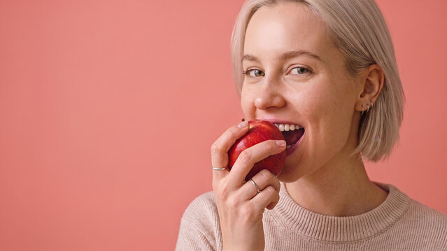A young, blonde woman in a knit sweater smiles while taking a bite of a red apple against a pink background.