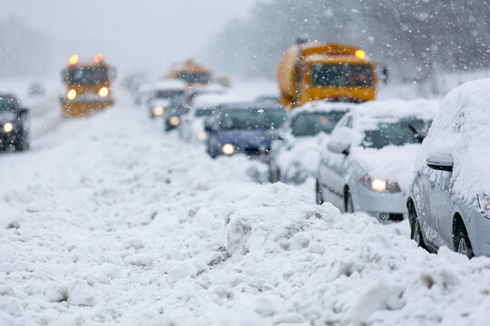 Snow blankets roads, turning streets into white, icy paths. Vehicles halt, lights dimmed as snowflakes drift downward. Yellow trucks wait patiently amid the quiet chaos