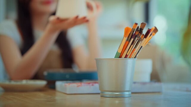 Paint brushes in metal container on table in ceramic pottery art studio workshop with Asian female artisan holding ceramic mug mug in background, blurred depth of field, creativity and small business 