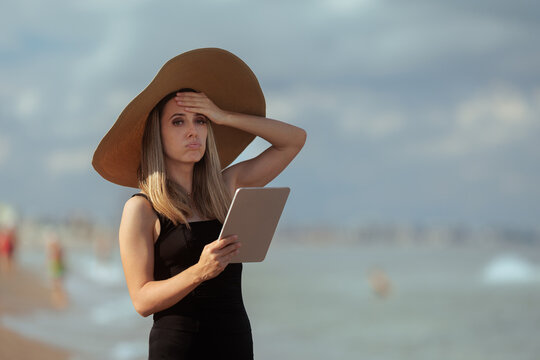 Stressed Woman In Large Hat Holding Tablet At The Beach. Anxious stressed female wearing swimwear holding digital device near ocean during summer vacation