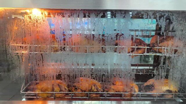 whole chickens being roasted slowly on a rotating spit inside a professional commercial rotisserie oven. The camera captures the golden-brown skin of the chickens glistening under the warm orange oven