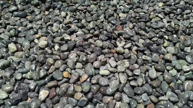water gently spraying over a bed of wet, dark pebble stones and black river rocks, creating a glistening, reflective texture. The steady stream of water droplets highlights the natural, smooth shapes 