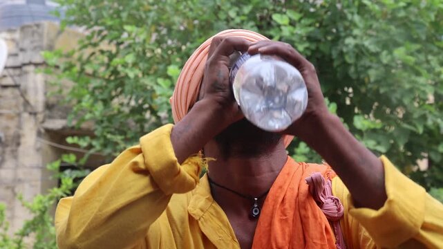 Indian Hindu sadhu drinking water from a plastic bottle outdoors, highlighting daily life, spirituality, hydration, and traditional culture in rural India.