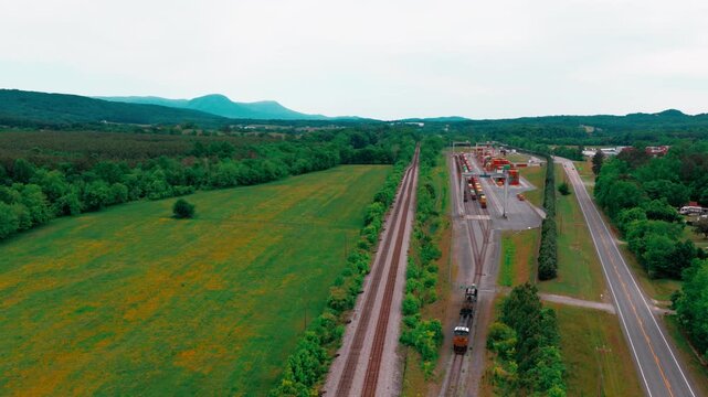 Rural intermodal rail terminal: gantry cranes over stacked containers as a freight train approaches on tracks beside pasture and Appalachian hills.